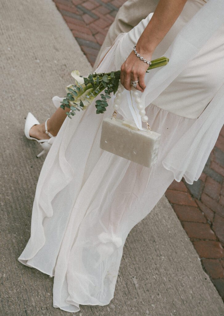 A bride's elopement outfit: a long white dress, white heels, a calla lily bouquet, and a pearl purse