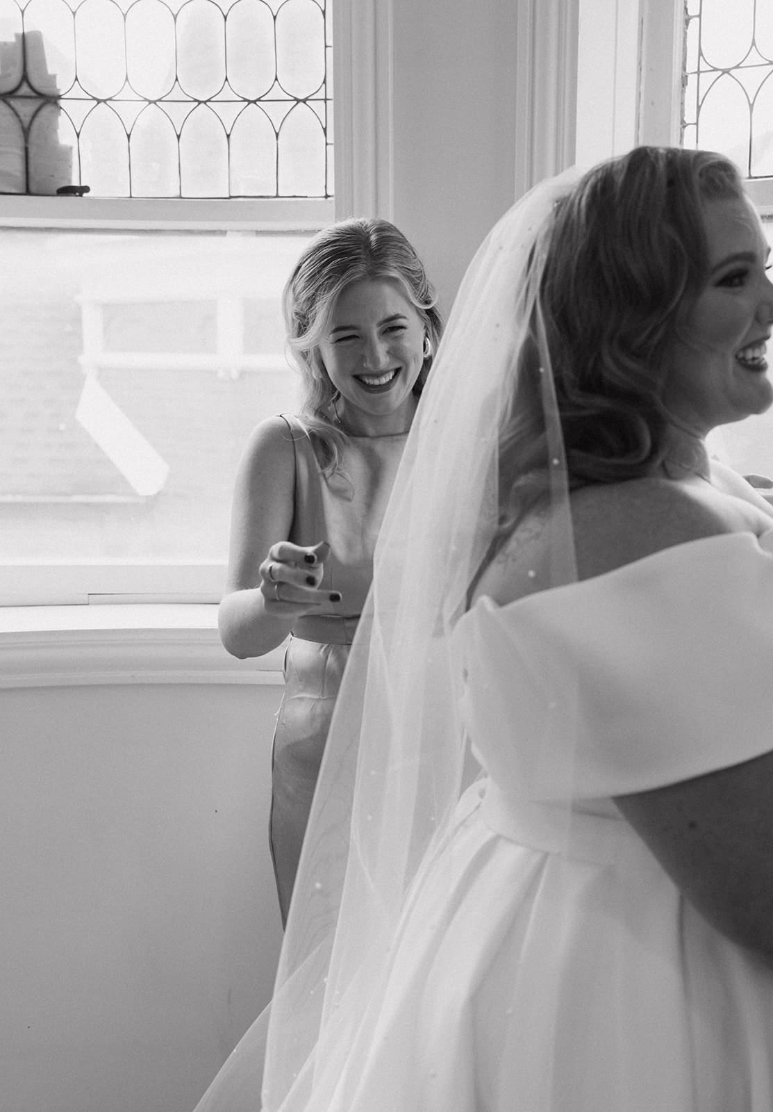 A bridesmaid laughing while helping put a veil on the bride